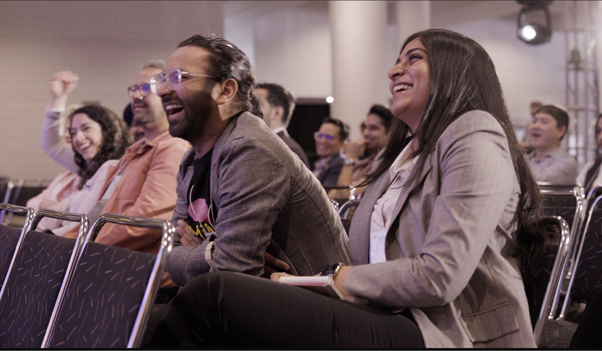 A group of people sit in rows of chairs at an indoor event, smiling and laughing as they watch something on stage. The atmosphere is lively and joyful, with everyone appearing engaged and entertained.